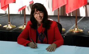 Dr. Cynthia Baker sits at a table signing a document. Canadian flags stand in the background.