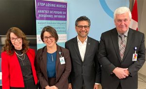 Four professionally dressed adults smile at the camera in front of a sign that reads “Stop losing nurses.” Dr. Cynthia Baker appears on the far left.