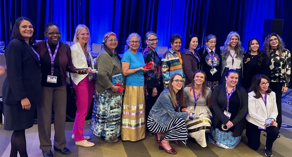 Lors d’une conférence, un grand groupe d’adultes portant des cordons porte-nom sourient à la caméra. Cynthia Baker, Ph. D. apparaît à l’extrême droite, au premier rang.